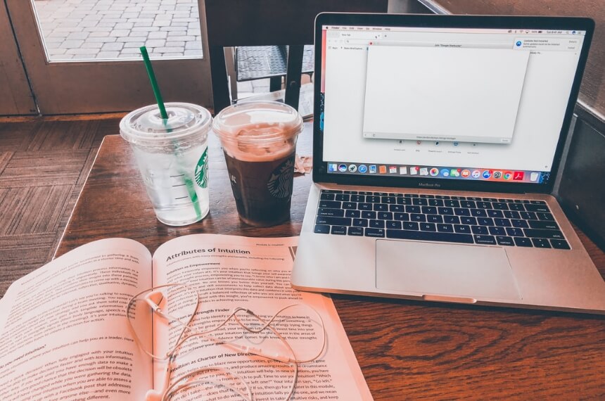 A laptop on a table with a Starbucks cup and an open book.