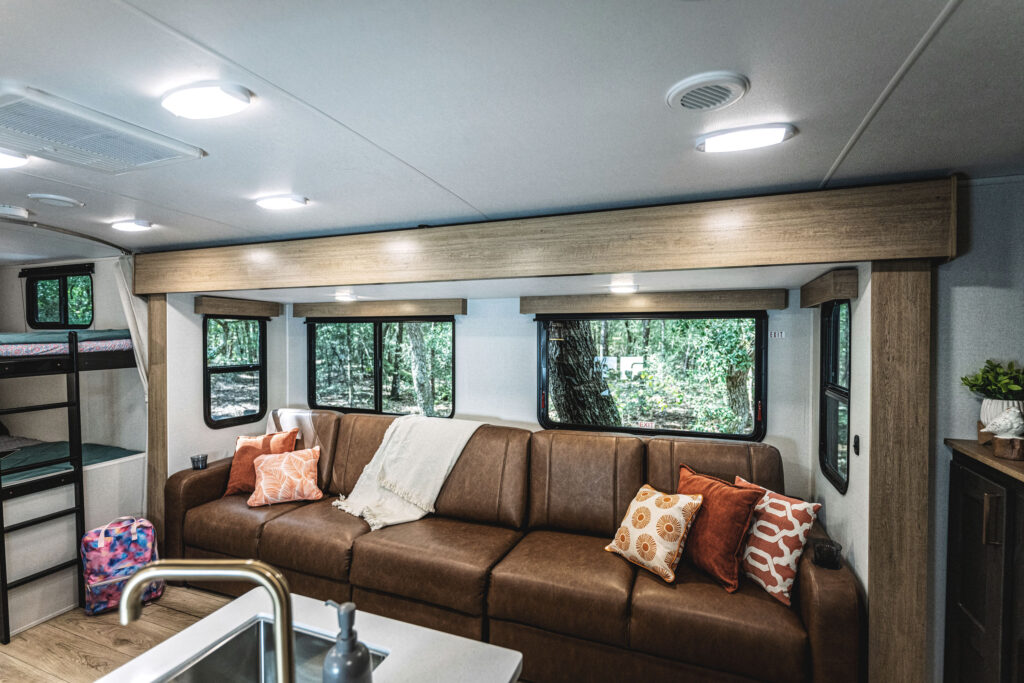 Interior of a modern RV featuring a brown leather sofa with colorful cushions, a kitchenette sink in the foreground, and large windows offering a view of the forest outside. Bunk beds and storage are visible in the background.