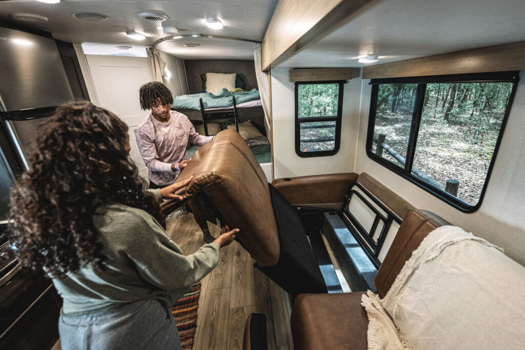 A man and woman are inside a camper van, lifting a couch cushion while preparing a bed. The van interior includes a kitchen area and a bunk bed, with windows showing a forest view outside.