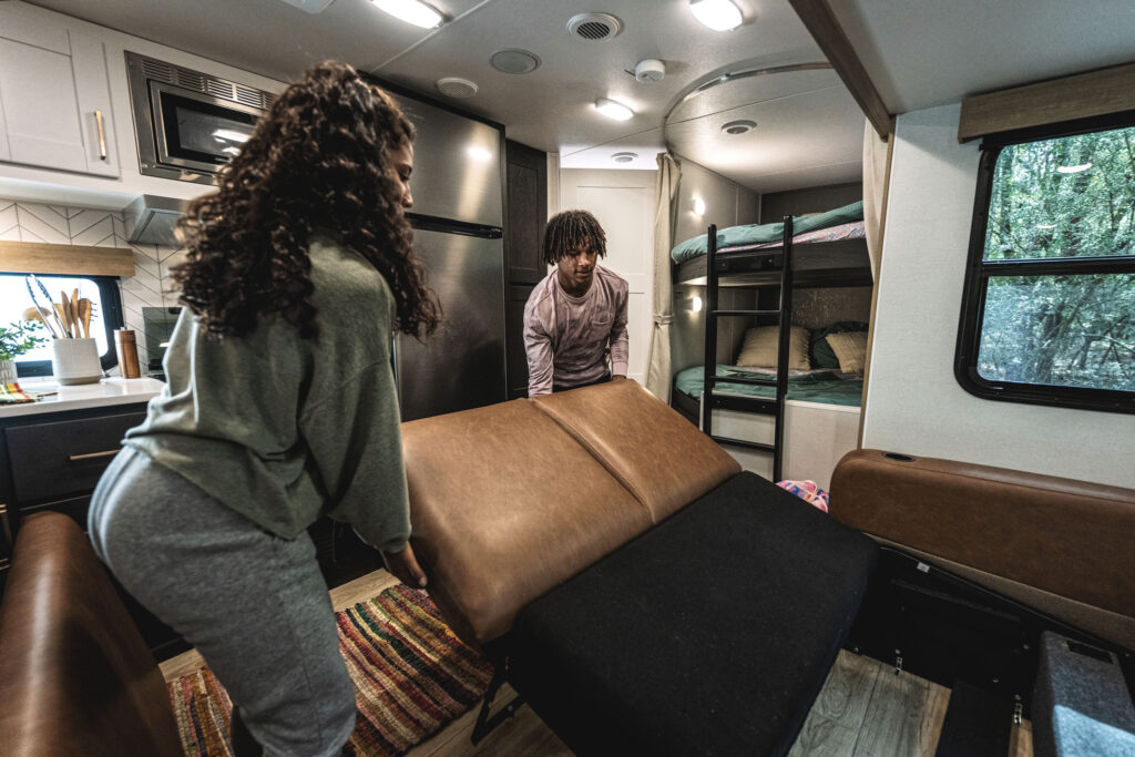 A woman and a man in comfortable clothing work together to unfold a sofa bed inside an RV. The interior is modern, featuring a kitchenette and bunk beds. A window shows trees outside. A colorful rug lies on the floor.