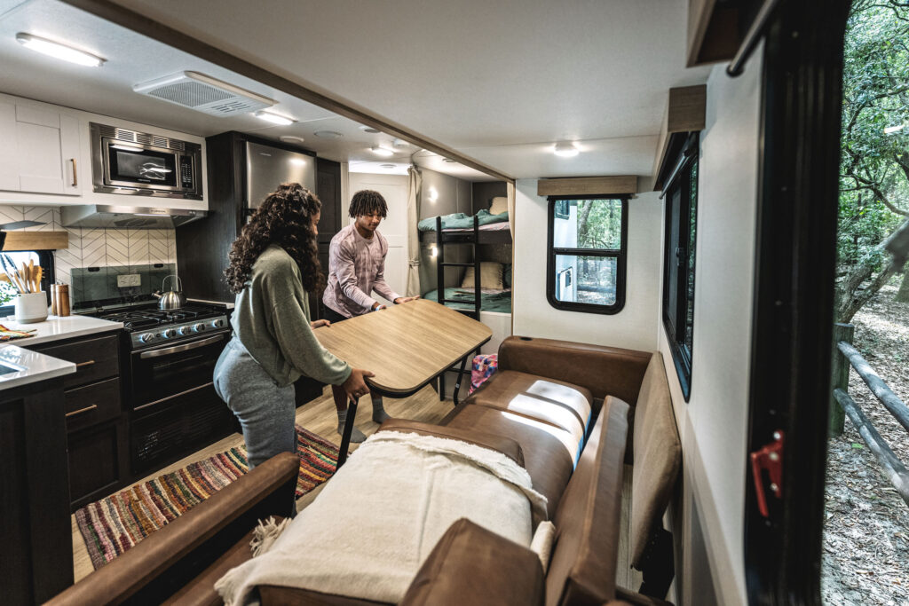 A man and woman are setting up a table inside a modern RV. The RV has a cozy interior with a kitchen area, a bunk bed, and a sofa. The window shows trees outside, suggesting they are in a wooded area.