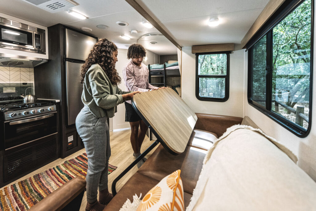 A couple adjusts a folding table inside an RV. The interior features a kitchen area with an oven and fridge, a colorful rug, and a couch. Large windows reveal a view of trees outside.