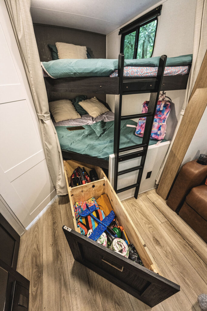 Bunk beds in a cozy room with a wooden floor. One bed has a colorful blanket, the other has a green one. A ladder provides access to the top bunk. Underneath, a drawer reveals various items, and a window brings in natural light.