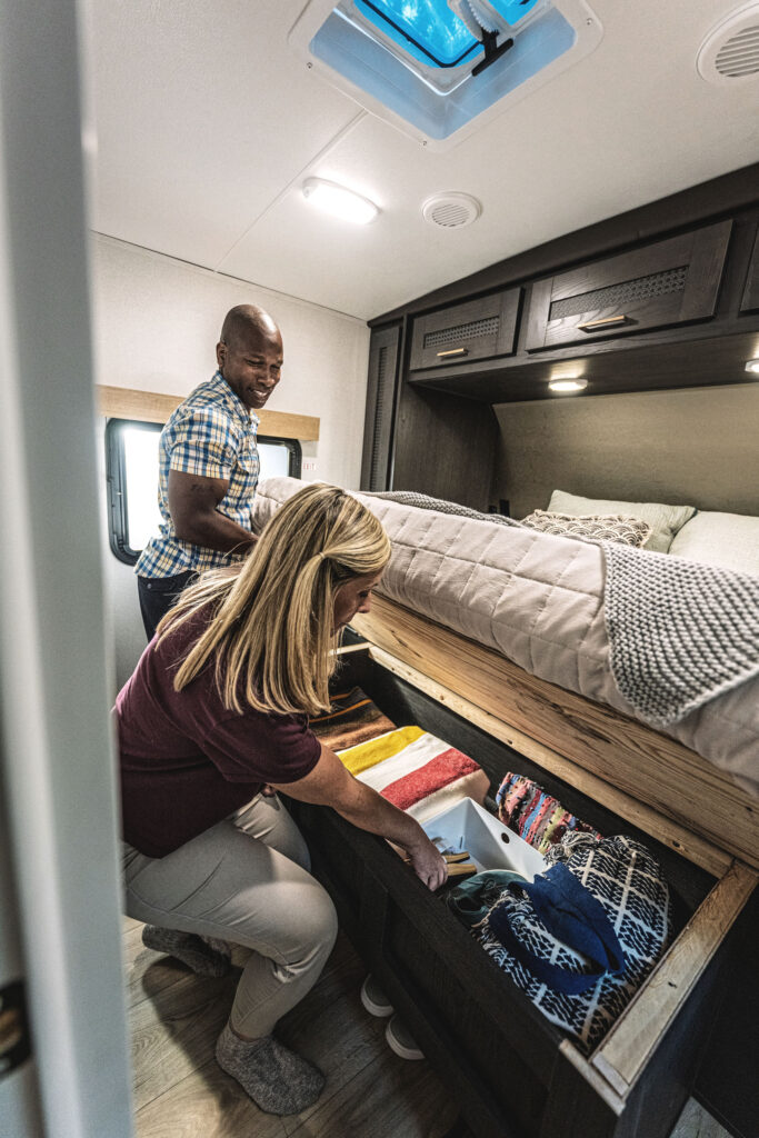 A man and a woman organizing a storage compartment under a bed inside a camper. She is placing folded clothes and towels neatly, while he watches. The interior is cozy with a window, overhead cabinets, and bedding in warm colors.