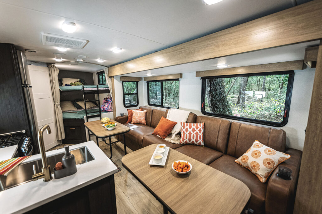 Interior of an RV featuring a cozy brown leather sofa with colorful cushions, a dining table with snacks, a kitchen area, and bunk beds in the background. Large windows offer a view of a wooded area outside.