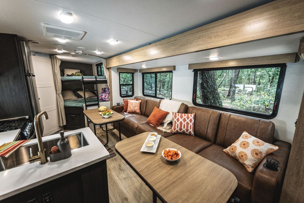 Interior of a camper featuring a kitchen sink, wooden cabinets, brown couch, and dining table set with snacks and drinks. Bunk beds are visible in the back, and large windows reveal a forested outdoor setting. Cozy and modern design.