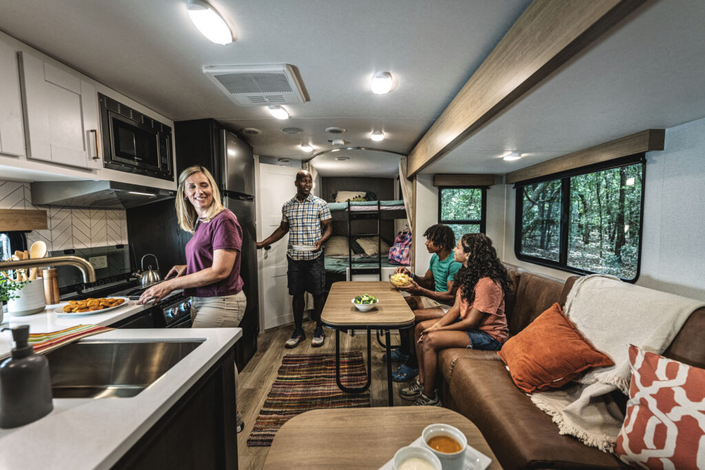 A family in an RV: a woman cooking at the kitchenette, a man standing near bunk beds, and two children sitting at a table. The RV has modern amenities with a cozy atmosphere, featuring a sink, stove, seating area, and lush green views outside the windows.