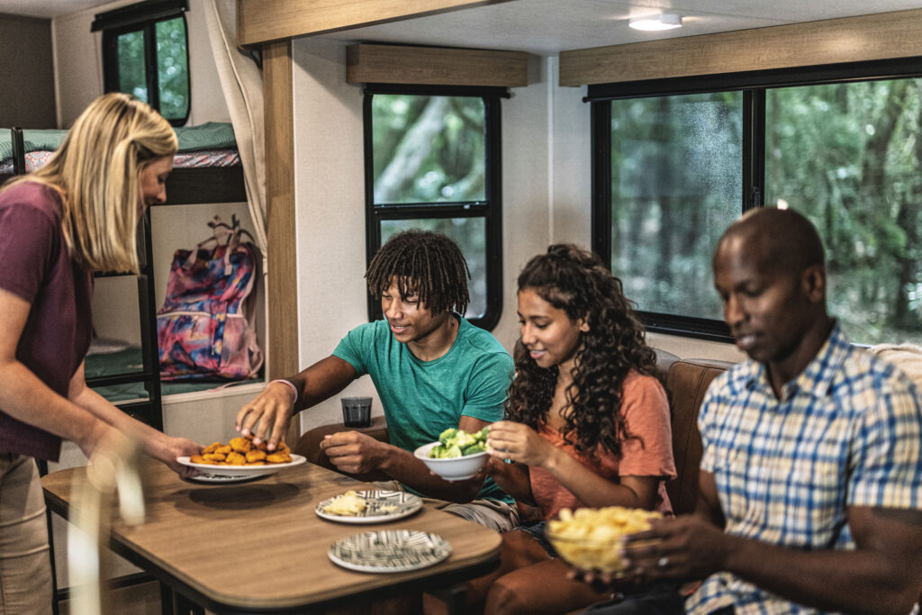 A group of four people is enjoying a meal inside an RV. Three people sit at a table with plates of salad, chips, and chicken nuggets. A woman stands beside them holding a dish. The interior is bright with large windows showing trees outside.