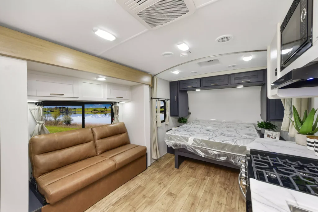 Interior of a modern RV featuring a brown leather sofa, a double bed with gray and white bedding, wood-style flooring, kitchen area with stove, microwave, and potted plants, with large windows overlooking nature.