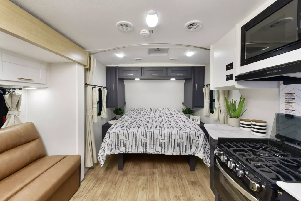 A modern RV interior features a bed with a geometric-patterned bedspread, overhead cabinets, a brown sofa, a kitchen area with a stove and microwave, potted plants, and wood-style flooring.