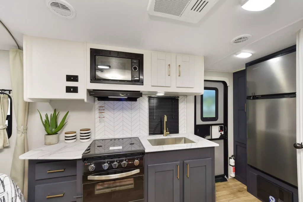 Modern RV kitchen with dark gray lower cabinets, white upper cabinets, a stainless steel sink, a stove with oven, a microwave, a large fridge, a potted plant, and a patterned backsplash. Bright and organized space.