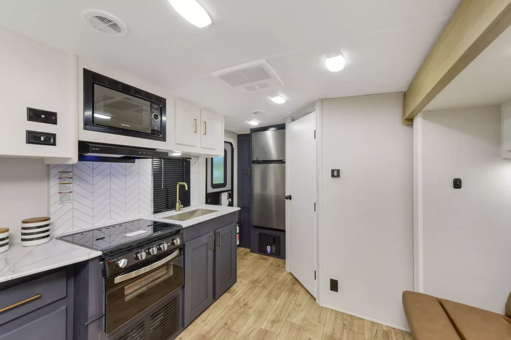Modern RV kitchen with a stove, oven, microwave, sink, and refrigerator. The space features light wood flooring, white and gray cabinetry, and a herringbone tile backsplash. Bright ceiling lights illuminate the area.