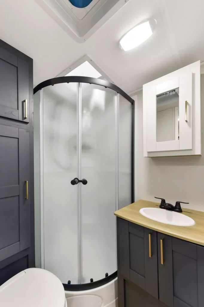 A modern, compact bathroom features a corner shower with frosted sliding doors, a gray vanity with a wood countertop, a black faucet, a wall-mounted mirrored cabinet, and a toilet partially visible.