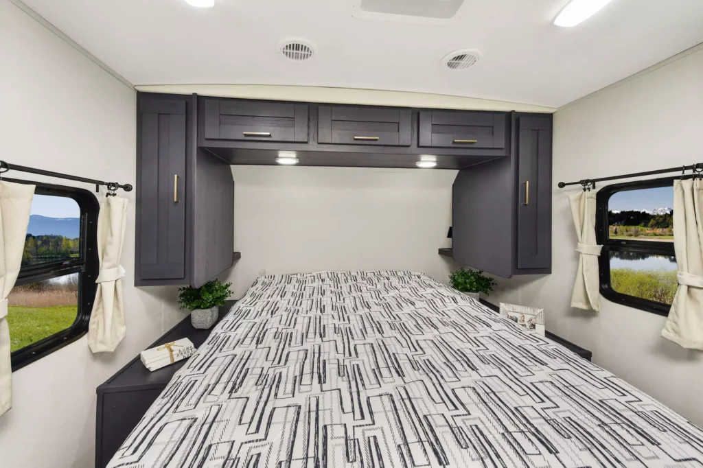 A neatly made bed with a geometric-patterned duvet sits between two windows in an RV bedroom. Overhead cabinets and nightstands with potted plants are on each side of the bed.