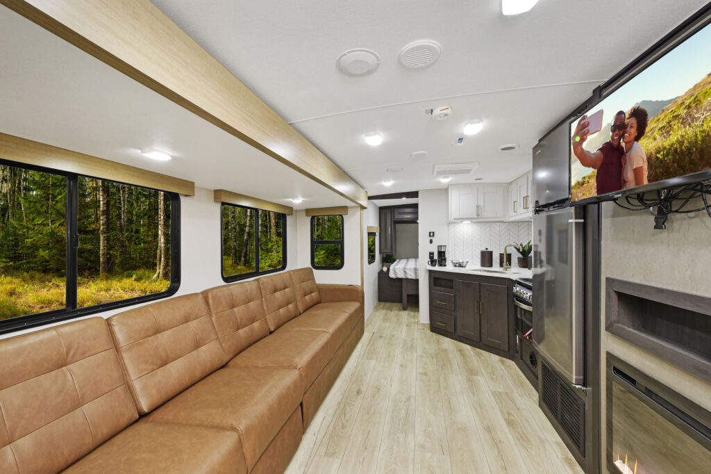 Inside this ultra-lightweight travel trailer, youll find a long brown leather couch, wood-look flooring, and a mounted TV. The kitchen area features a countertop, sink, and cabinets. Large windows offer stunning views of the forest outside.