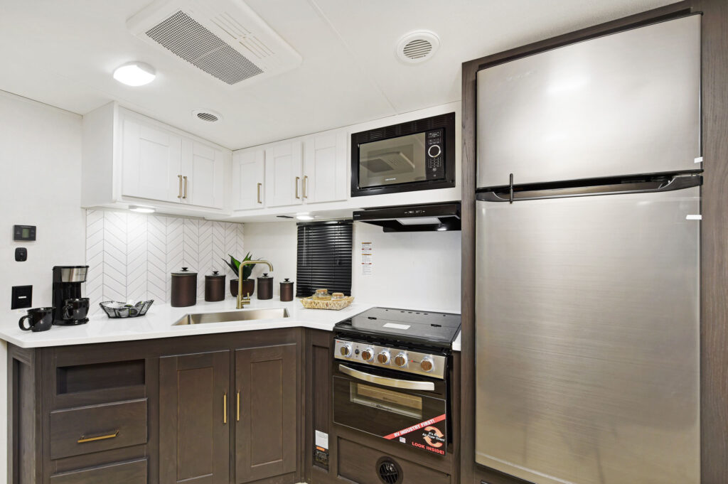 Modern RV kitchen interior in ultra-lightweight travel trailers features a stainless steel fridge, oven, and microwave. White cabinets above, darker ones below. A countertop hosts a coffee maker, plant, and containers. Chevron-patterned backsplash adds a stylish touch.