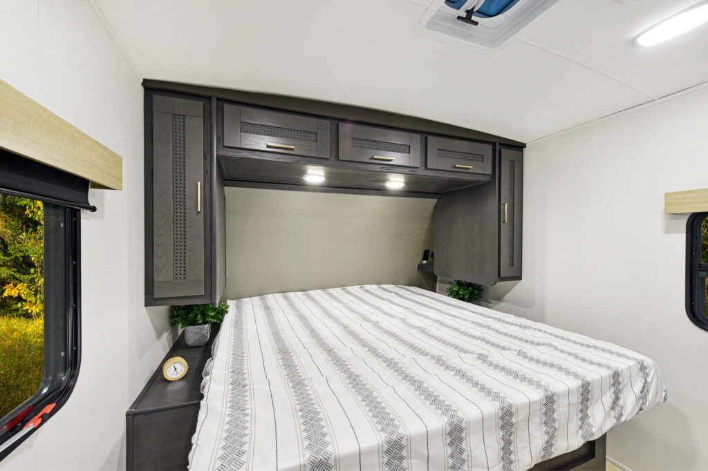 Inside this ultra-lightweight travel trailers bedroom, a neatly made bed with a striped duvet beckons. Above the bed, dark wood cabinets with built-in lights offer elegance. Natural light pours in through a window on the right, while a small plant and clock adorn the bedside shelf.