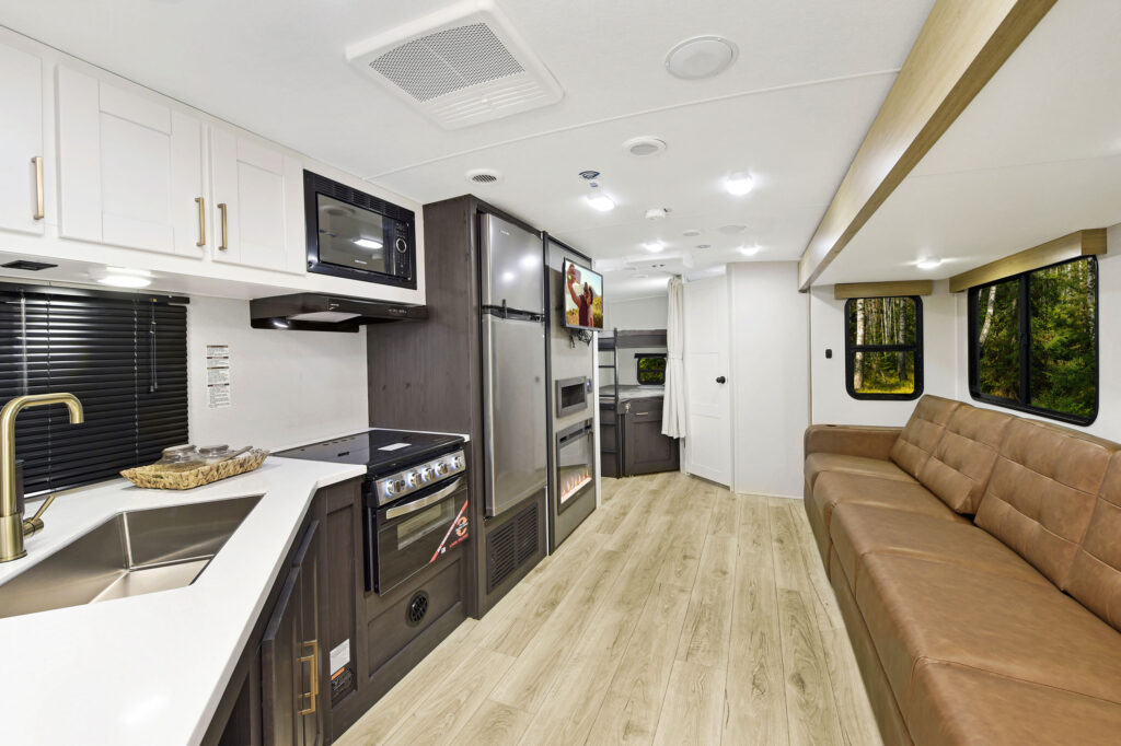 The interior of this ultra-lightweight travel trailer showcases a modern RV design, featuring a kitchen with white cabinets, black microwave, and oven on the left. On the right is a brown leather couch atop light wood flooring. A flat-screen TV is mounted near a quaint dining area at the back.