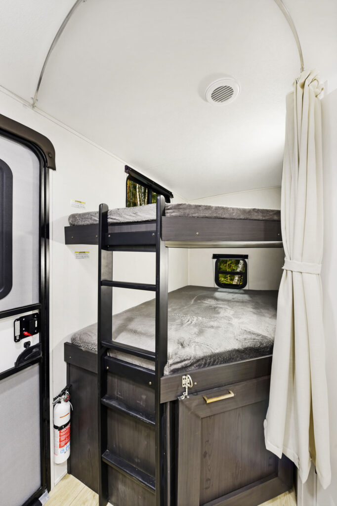 Interior of an ultra-lightweight travel trailer showcasing a double-decker bed setup. The lower bunk features a window, while the upper bunk has a small horizontal one. A black ladder and white curtain add charm, with a fire extinguisher conveniently nearby for safety.