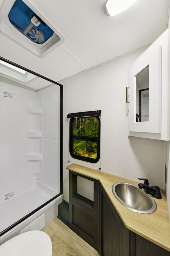 The interior of this ultra-lightweight travel trailers bathroom features a shower on the left, a window overlooking greenery, and on the right, a vanity with a round sink, faucet, and mirrored cabinet—bringing comfort and style to your adventures.
