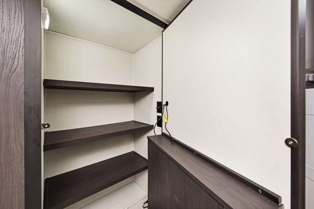 Inside view of an open, empty closet in a sleek ultra-lightweight travel trailer, featuring dark wooden shelves and a white interior. The closet door is partially open, revealing two horizontal shelves and an electrical cable attached to the side.