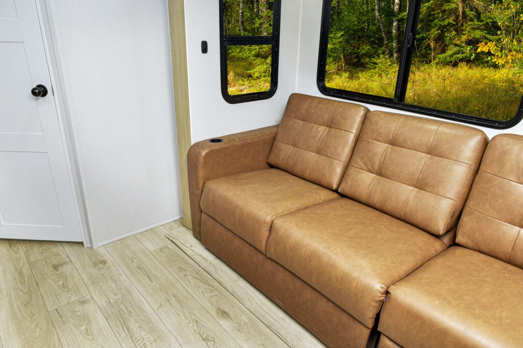 The interior of an ultra-lightweight travel trailer features a brown leather couch against a white wall with a black-framed window. Outside, lush forest greenery is visible. The floor is light wood, with a partially seen door on the left, blending comfort and nature seamlessly.