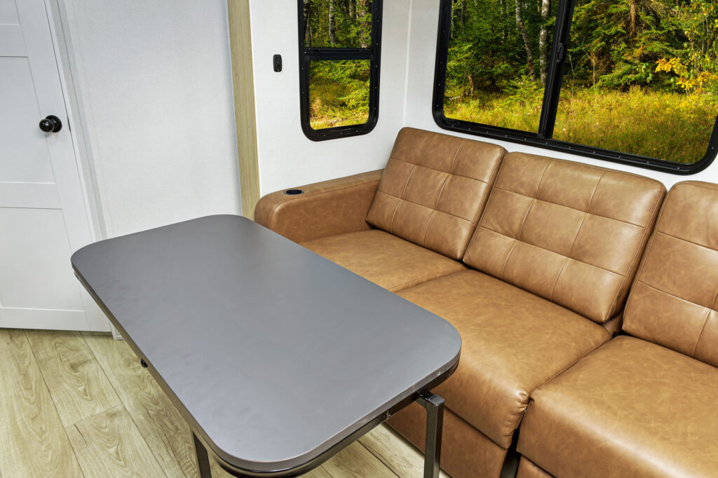 Interior of a modern, ultra-lightweight travel trailer featuring a brown leather sofa and a folding gray table. Large windows reveal a view of a green, wooded area outside. White walls and light wood flooring create a bright and airy atmosphere.