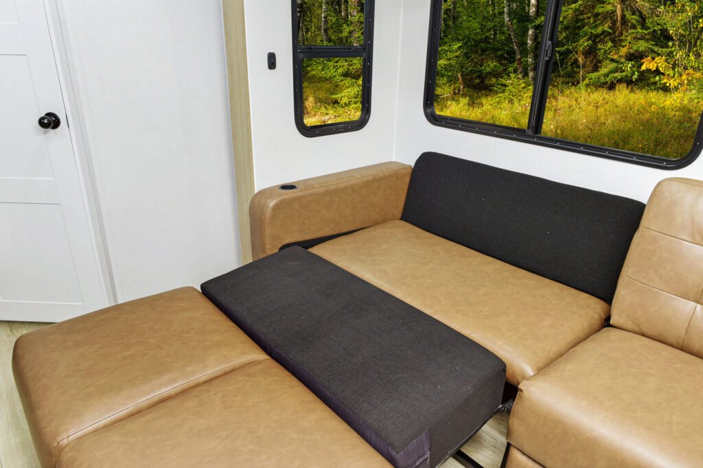The ultra-lightweight travel trailers interior boasts a convertible sofa bed with tan cushions and a black mattress, positioned near windows offering serene forest views. Bright natural light beautifully highlights the compact living space.