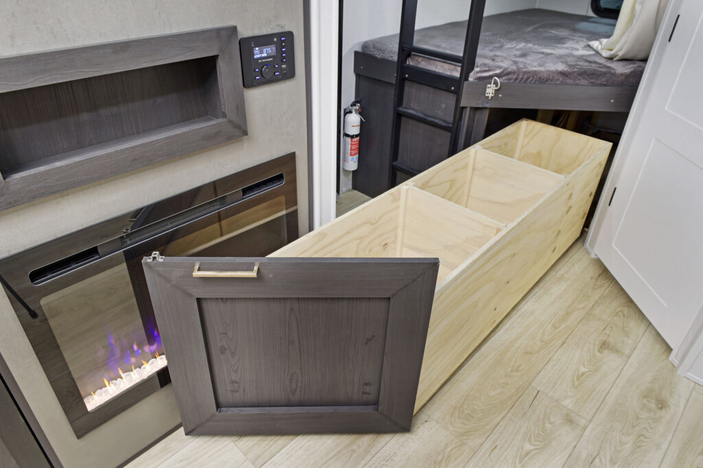 Inside an ultra-lightweight travel trailer, youll find an open wooden storage compartment with a faux fireplace and control panel adorning the wall. A cozy bunk bed and fire extinguisher sit nearby, all set against a backdrop of light-colored wood flooring.