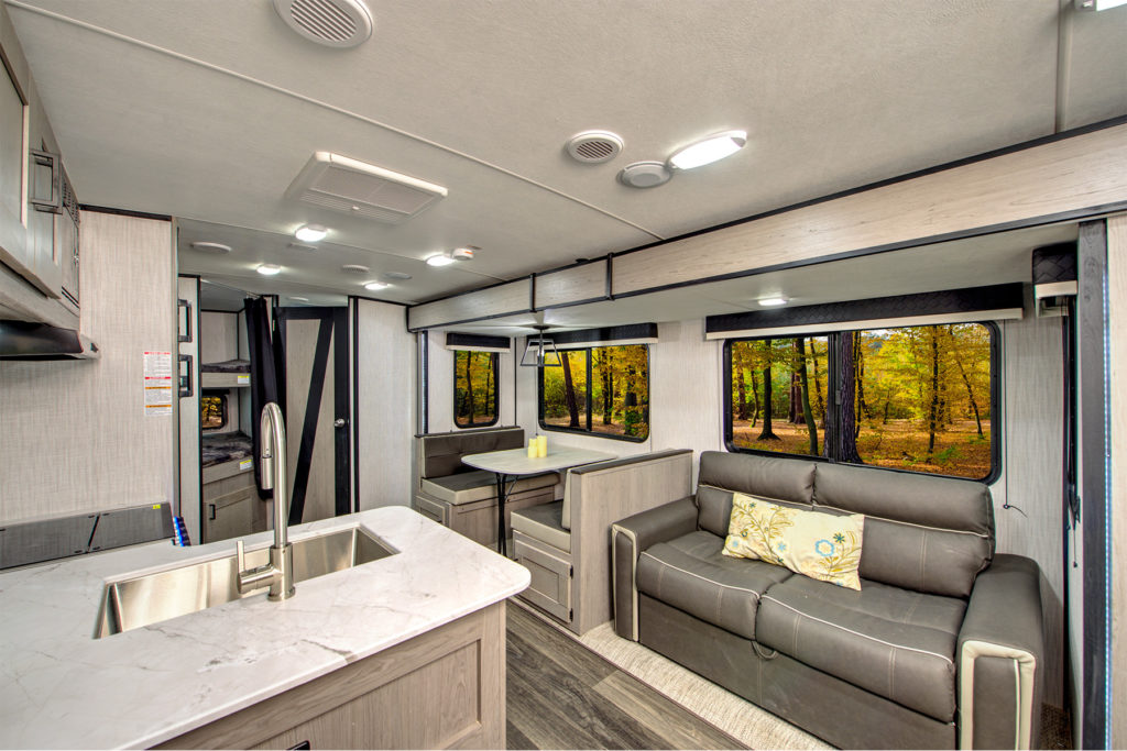 Modern RV interior with a marble countertop kitchen, dining booth, gray sofa with a patterned pillow, large windows showing a forest scene, and bright overhead lighting.