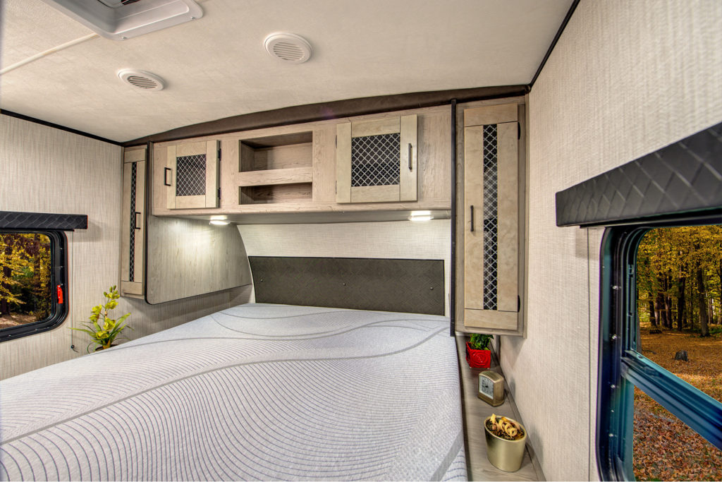 A cozy RV bedroom features a neatly made bed with a light gray patterned comforter, overhead cabinets, small decor items on the shelves, and a window showing a forest with autumn foliage.