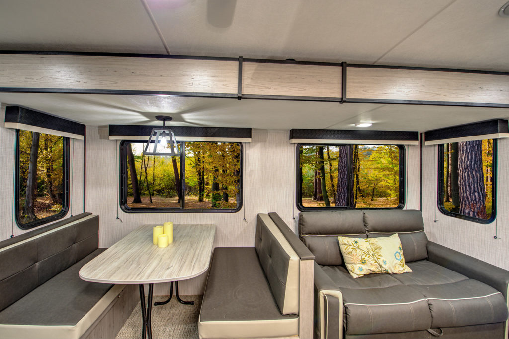 Interior of a modern RV with a gray and beige dinette set, a sofa with decorative pillows, large windows showing a forest with autumn foliage, and a ceiling light above the table.