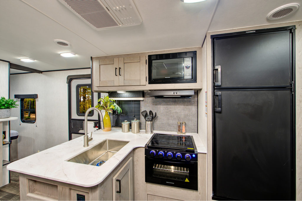 Modern RV kitchen with a marble countertop, stainless steel sink, electric stove, microwave, and a black refrigerator. There are cabinets, cooking utensils, and a plant in a yellow vase on the counter.