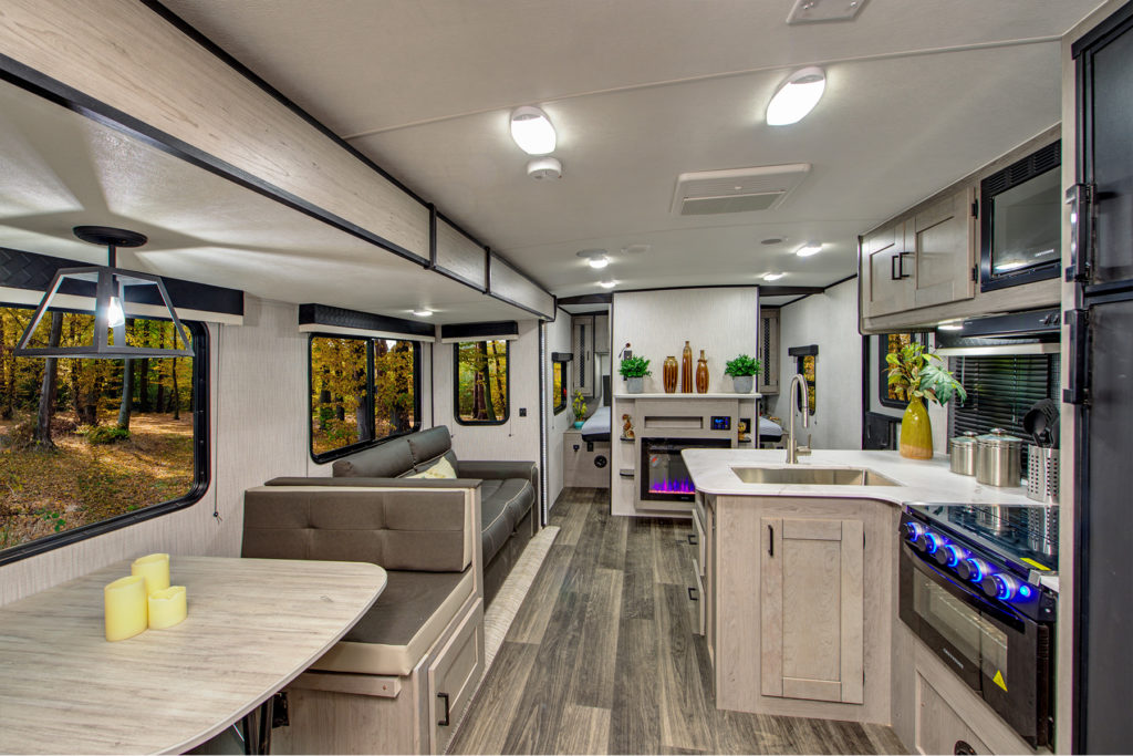 Modern RV interior featuring a kitchen, dining table, sofa, and electric fireplace. Large windows show a forest view. Light wood cabinets, gray seating, and decorative plants create a cozy, spacious atmosphere.