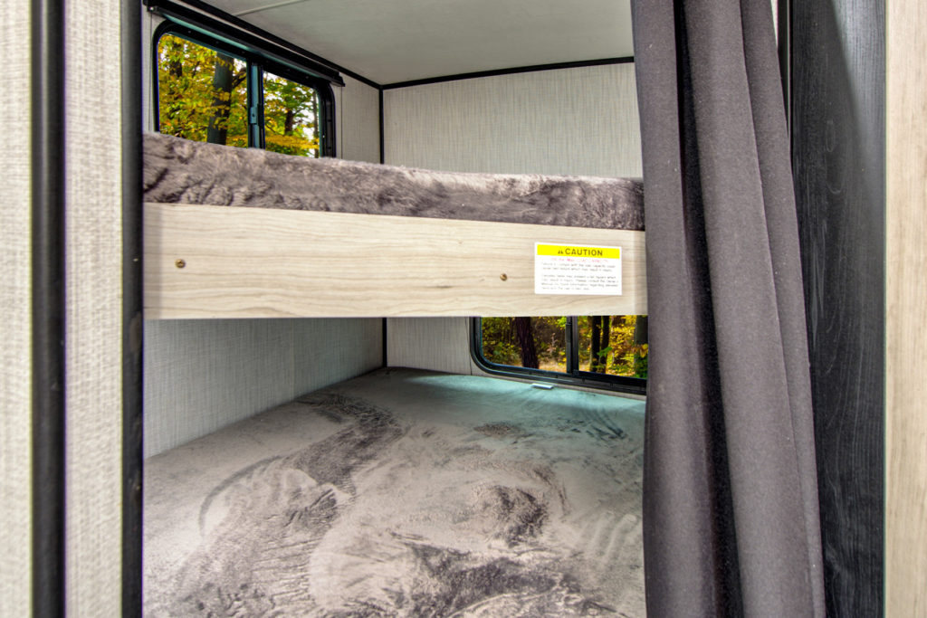 Bunk beds inside an RV with gray bedding, a window next to each bed, a curtain partially covering the entrance, and a caution sign on the top bunks frame. Trees are visible outside the windows.
