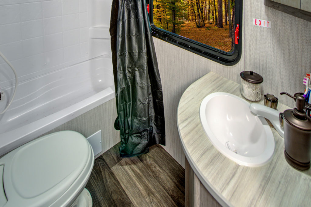 Small RV bathroom with a toilet, white sink on a gray countertop, and a bathtub with a black shower curtain. A window shows a forest scene with sunlight streaming through trees.