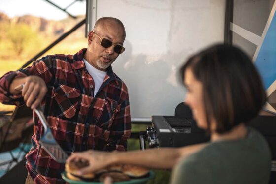 A family cooking outdoors beside the 2024 Heartland Cyclone 4008 Fifth Wheel Toy Hauler, enjoying a barbecue under a clear sky.