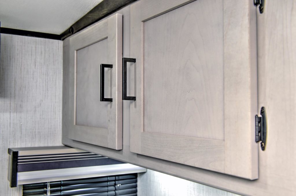 Light wood kitchen cabinets with black handles are mounted on a wall above a window with black blinds and a valance. The wall has a subtle textured pattern.