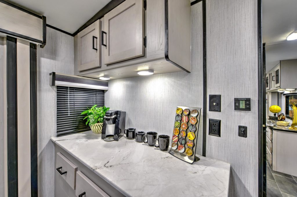 A modern RV kitchen counter with a marble surface, black mugs, a Keurig coffee maker, a coffee pod holder, a potted plant, and cabinets above, all under bright lighting.
