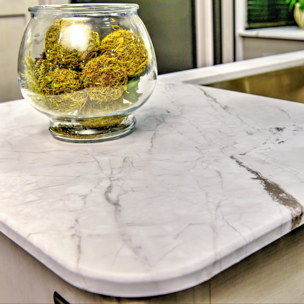 A round glass bowl filled with green moss sits on a white marble countertop with subtle gray veining and a polished, rounded edge.