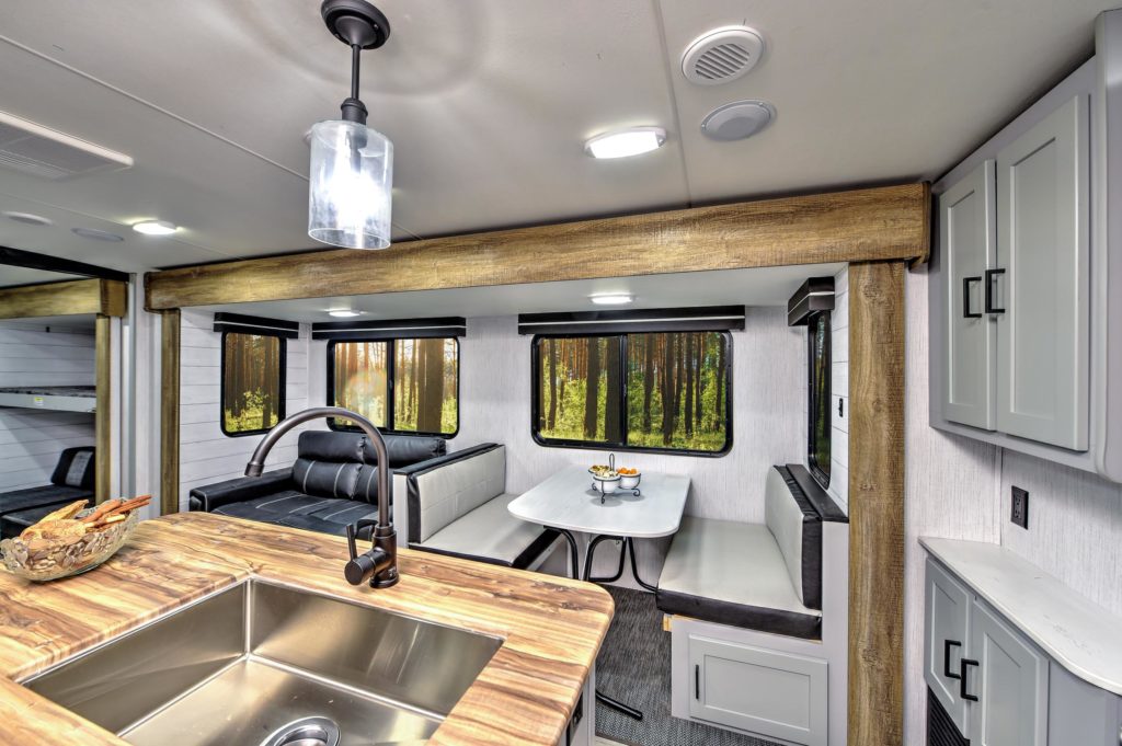 Modern RV interior showing a kitchen with a wood countertop and sink, and a dining area with a white table, black-and-white cushioned benches, large windows, and light grey cabinetry. Forest scenery is visible through the windows.