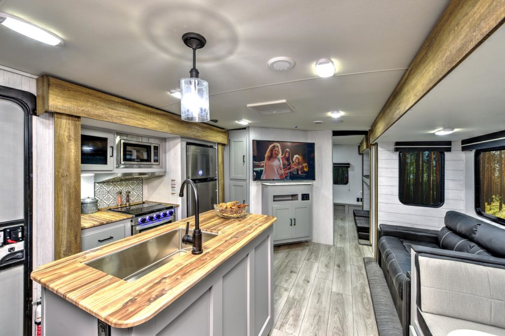 Modern RV interior with a wood-accented kitchen, stainless steel appliances, and a cozy living area featuring a TV and large windows overlooking a forested view. Light wood flooring and stylish decor create an inviting atmosphere.