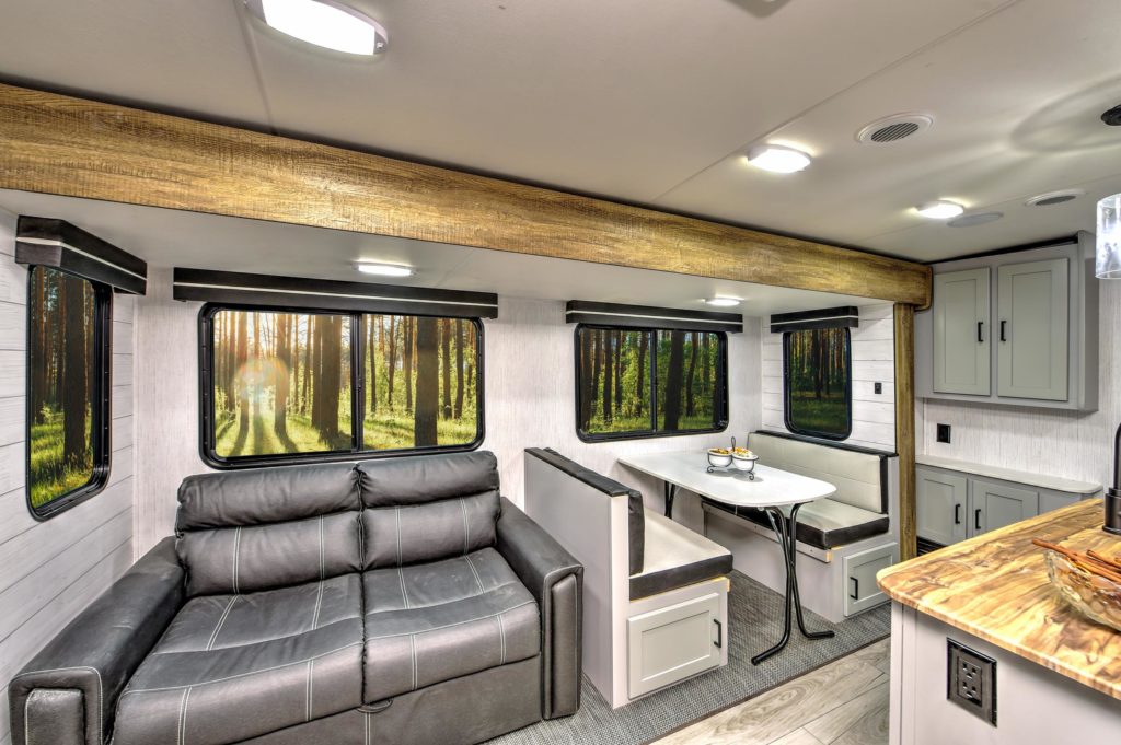 Interior of a modern RV with a black leather couch, a white dining table with bench seating, large windows showing a forest view, and a compact kitchen with a wood-style countertop.