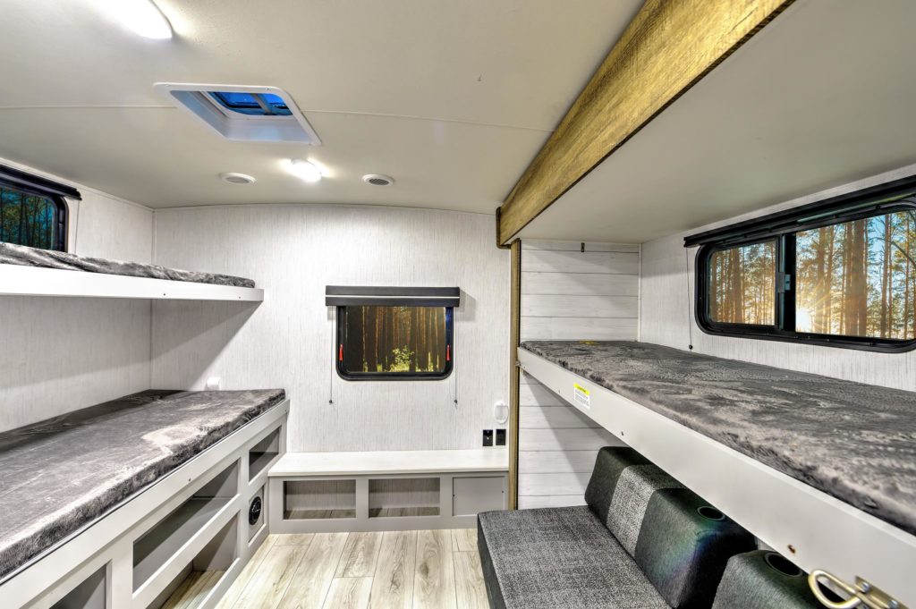 Modern RV interior with two sets of bunk beds, grey cushions, wood-look flooring, large windows, and built-in storage. Sunlight streams through forest trees visible outside the windows.
