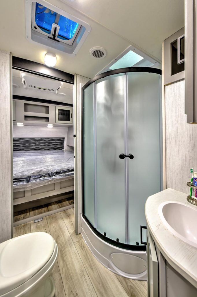 Modern RV bathroom with a curved frosted glass shower, white toilet, small sink with toiletries, and a view into a bedroom with a made bed and overhead lights. Light wood flooring and bright, clean decor.