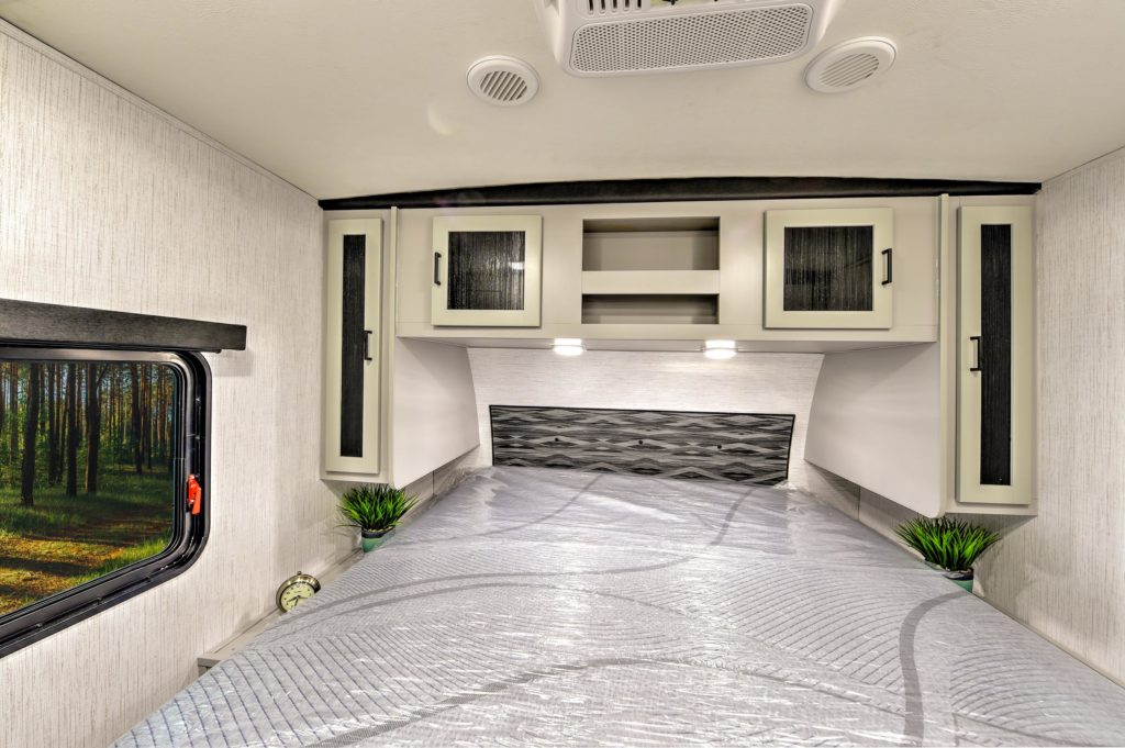 Interior of a camper bedroom with a covered mattress, built-in overhead cabinets, two small potted plants on each side, and a window showing a forest view. Ceiling lights and air vents are visible.