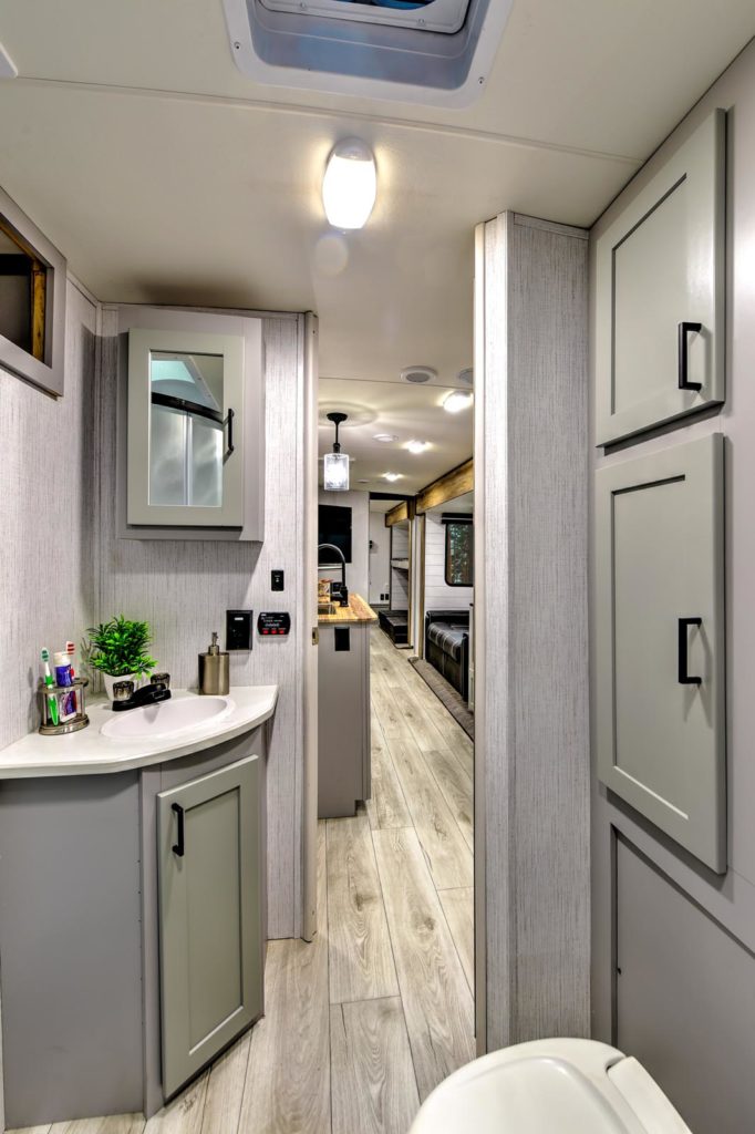 Modern RV bathroom with a small sink, vanity, mirror, storage cabinets, and toiletries visible. The hallway leads to a well-lit kitchen and living area with wood-like flooring and contemporary decor.