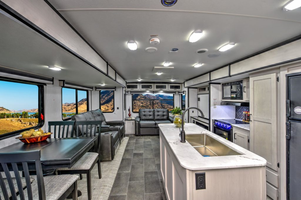 Modern RV interior with a kitchen island, stainless steel sink, dining table, black sofas, and large windows showing mountains and landscape outside. Bright lighting and sleek, neutral décor throughout.
