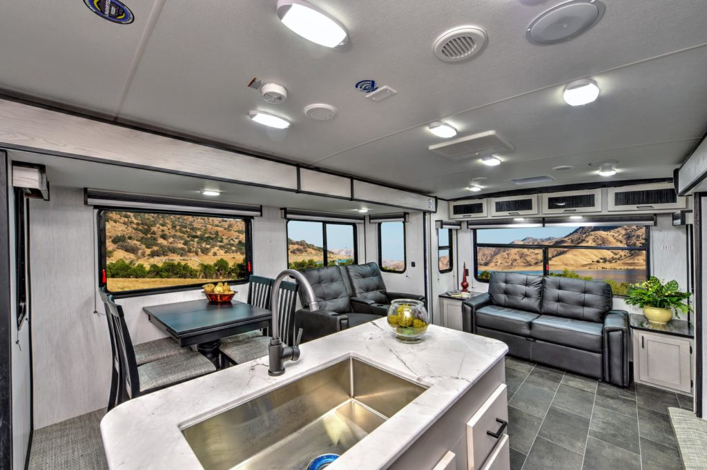 Modern RV interior with marble countertop, stainless steel sink, gray sofas, dining area, large windows showing scenic mountains and hills, and bright, well-lit space with overhead lighting and contemporary decor.