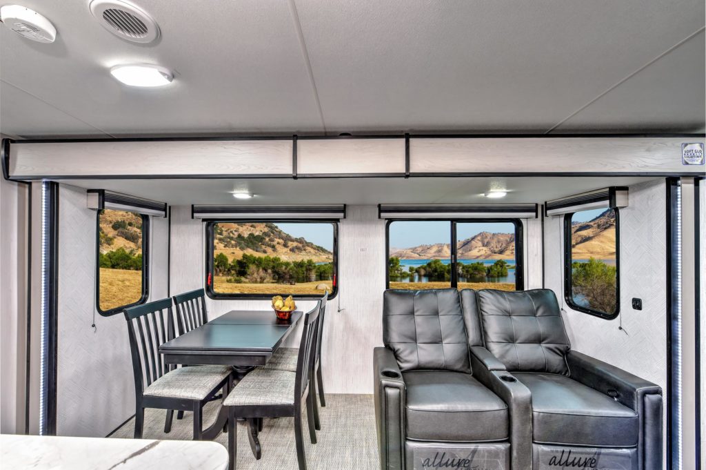 A modern RV interior features a dining table with benches and a bowl of fruit on the left, and two gray recliners on the right. Large windows reveal a scenic view of hills and grassland outside.
